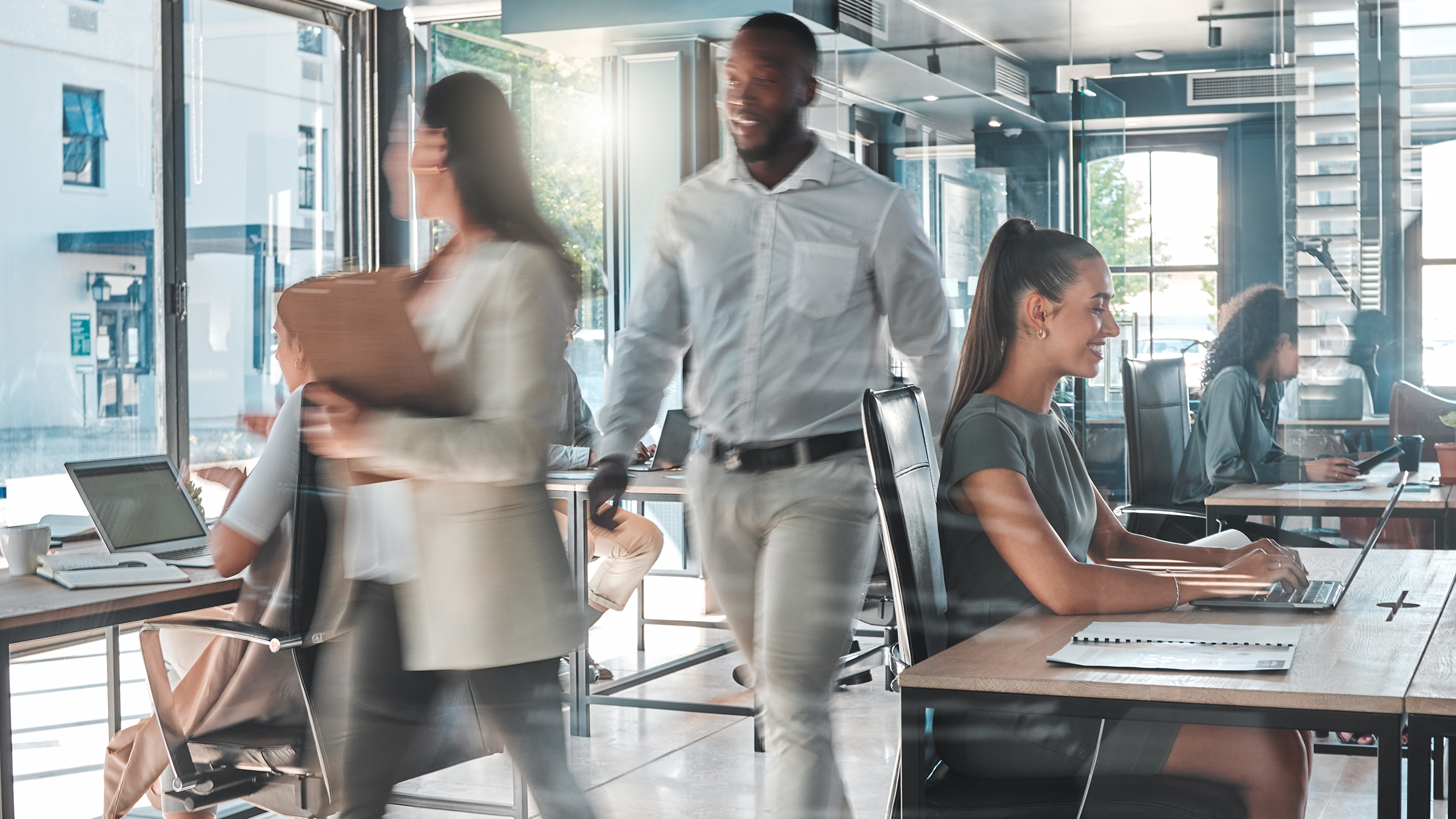 A contemporary office space featuring professionals engaged in various tasks. The setting includes large windows allowing natural light, desks with laptops, and a collaborative atmosphere. Individuals are seen walking and seated, creating a dynamic work environment. The scene highlights productivity and modern workplace design.