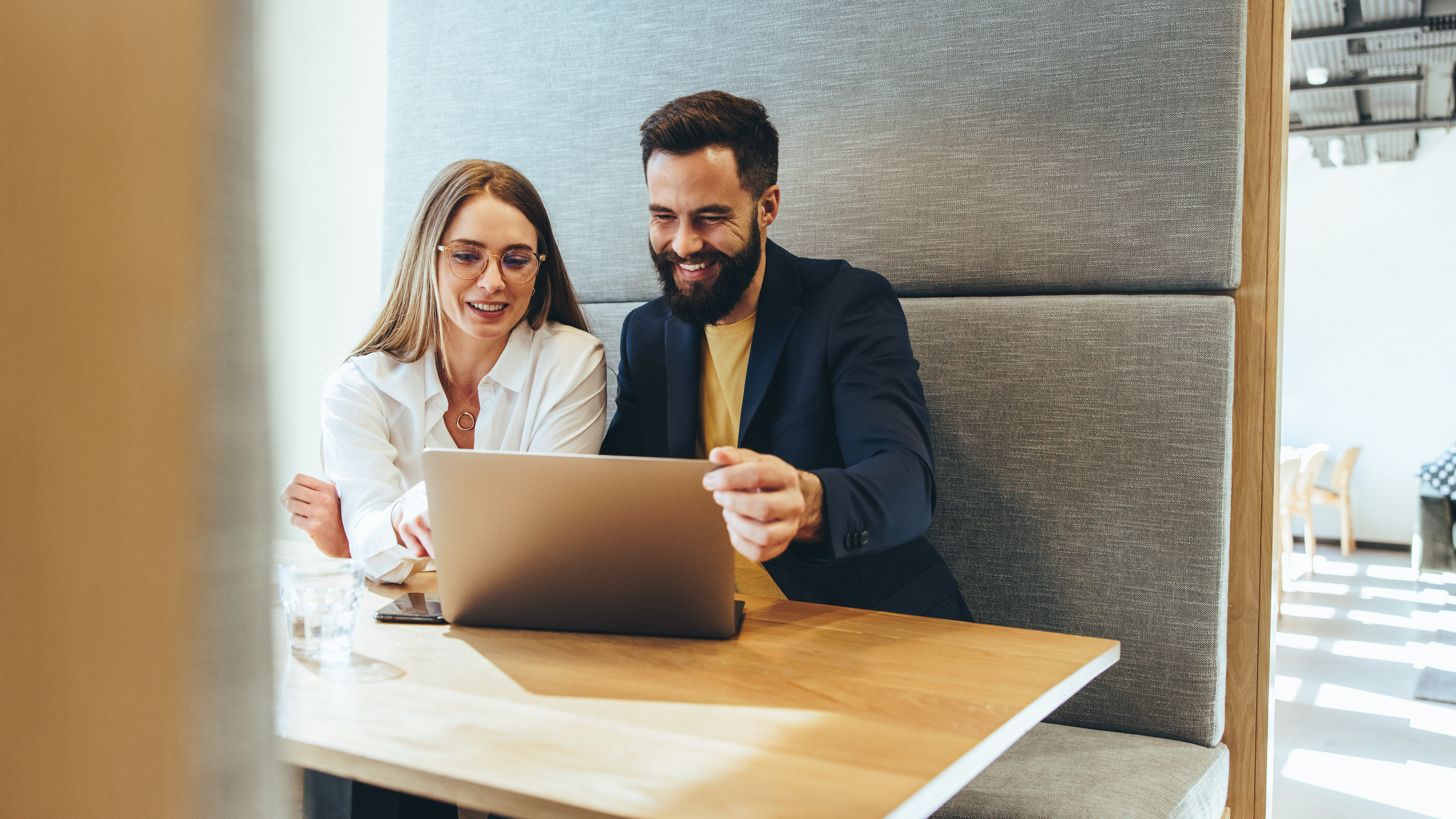 Two business professionals smiling at a laptop screen cheerfully. Happy young businesspeople working together in a modern workspace. Two successful young entrepreneurs collaborating on a new project.