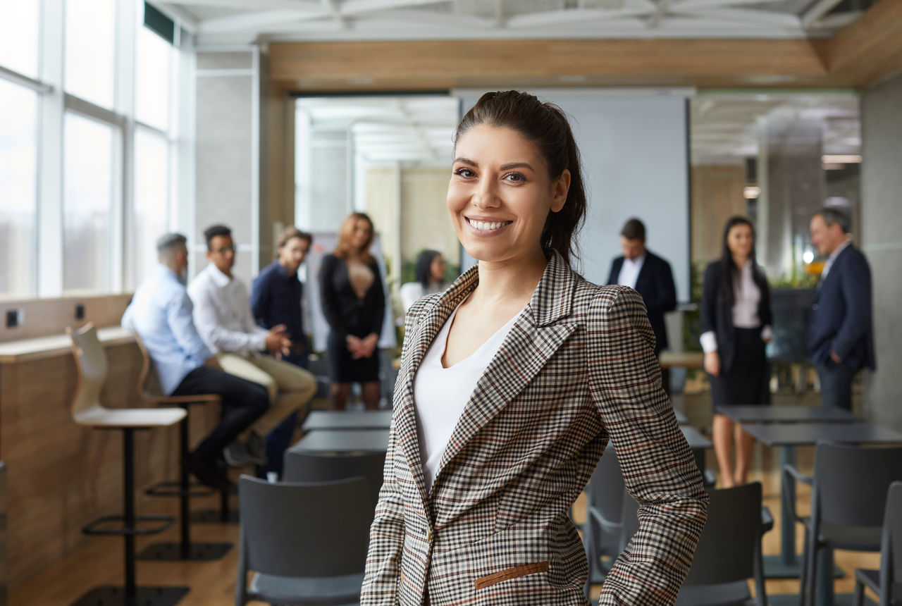 Portrait of happy female business teacher and professional coach. Beautiful young woman in jacket standing in office after corporate training class for team of employees, looking at camera and smiling